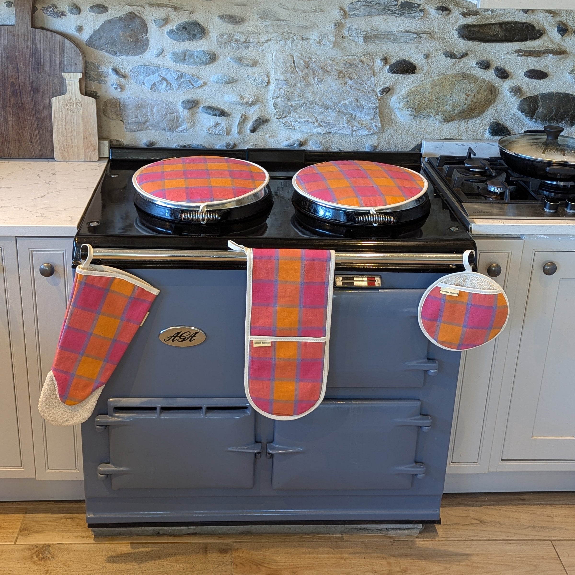 Blue Aga range cooker with checkered tartan like orange pink and blue pot holders and oven mitts and lid covers in a kitchen setting.