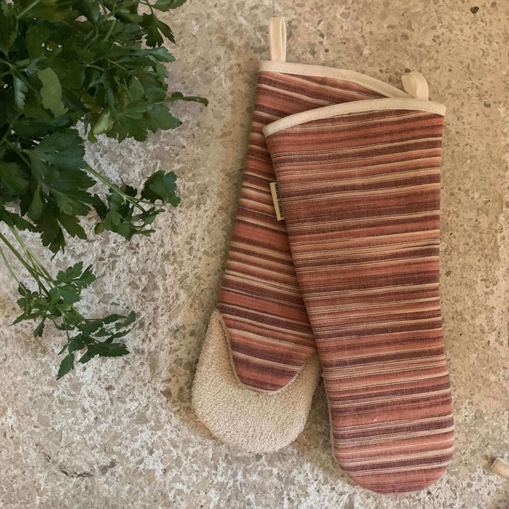 a pair of long oven gloves with red horizontal stripes on an off white marble top with fresh cooking herbs on the top left, flat image