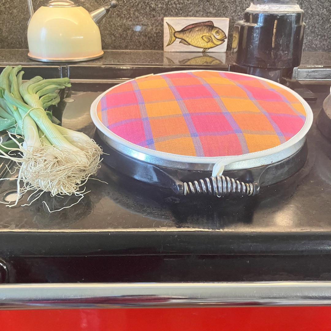 Colourful plaid Aga lid cover  pad on a stove with a white kettle and green onions in the background.
