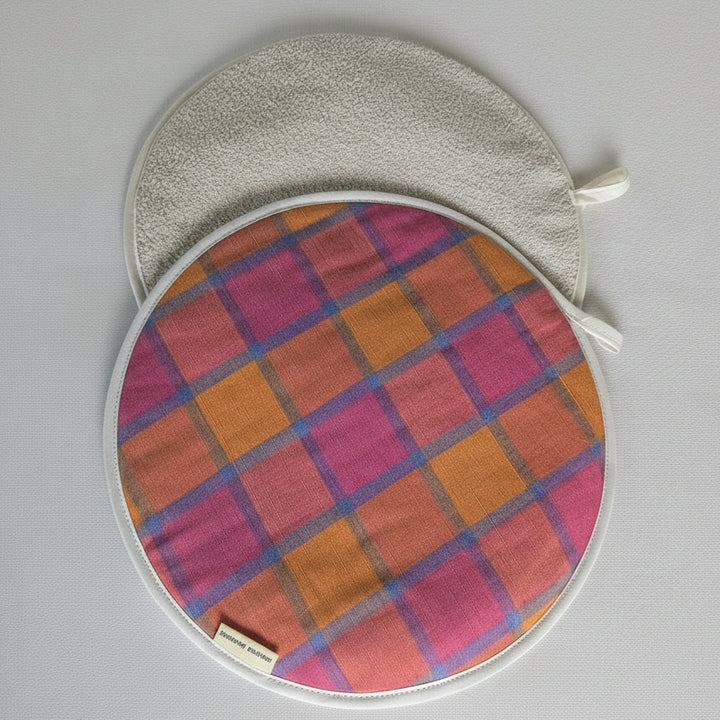 Two round fabric potholders with white borders, one featuring a pink, orange, and purple plaid pattern and the other a solid beige textured side, displayed on a light gray background.
