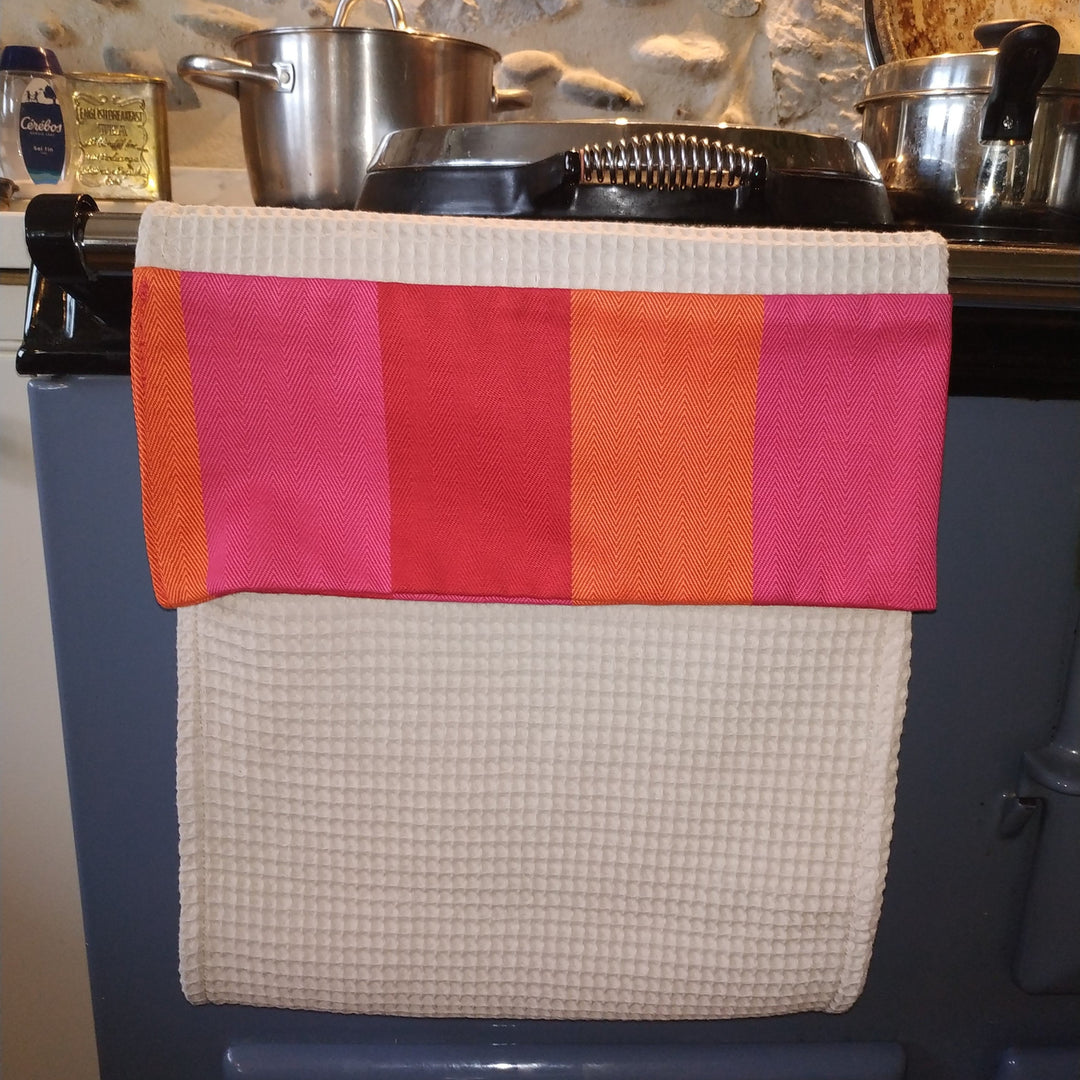A kitchen towel with a cream waffle-weave lower section and a striped top band in orange, pink, and red, hanging over a dark blue stove in a rustic kitchen setting.