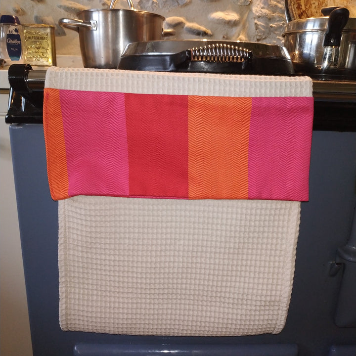 A kitchen towel with a cream waffle-weave lower section and a striped top band in orange, pink, and red, hanging over a dark blue stove in a rustic kitchen setting.