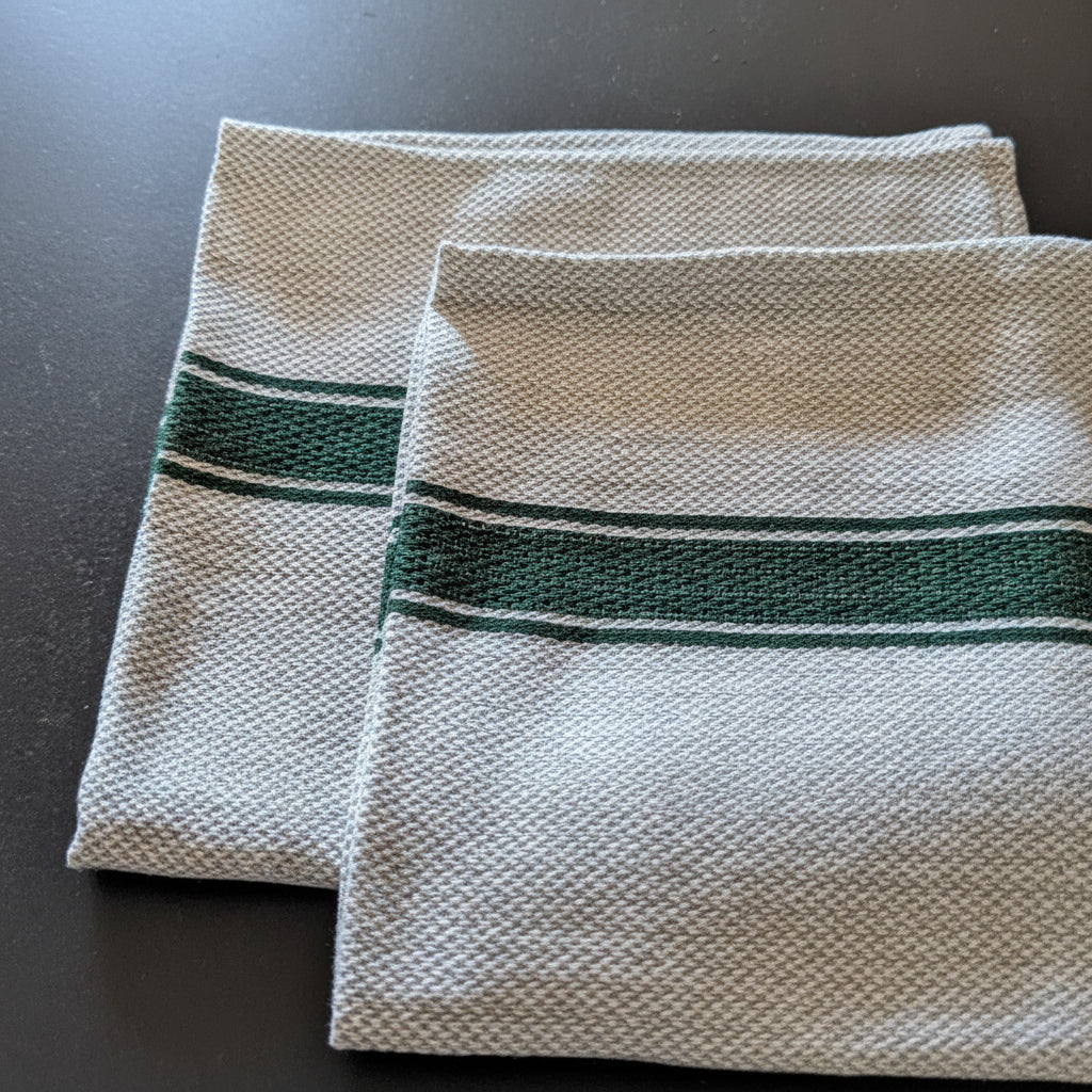 Two folded woven linen  kitchen towels with a dark green stripe detail, displayed on a dark gray surface.