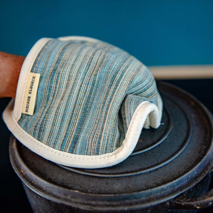 Close up of a pot holder blue with thin stripes with a person hand in it, lifting a the lid of a pot with a blue background