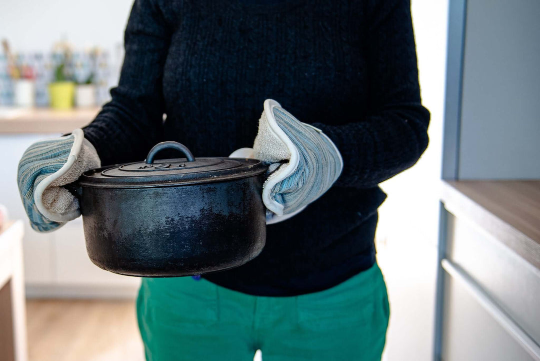 person with green trousers and black jumper close up of the middle holding a cast iron black pot with 2 blue striped pot holders oven mitts