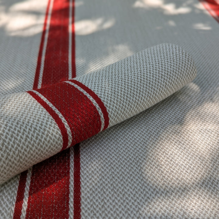 A close-up of a woven linen tablecloth with a light beige base, featuring bold red vertical stripes and thin white accent lines. A matching rolled napkin is placed diagonally across the surface, which is softly lit by dappled sunlight and shadows.