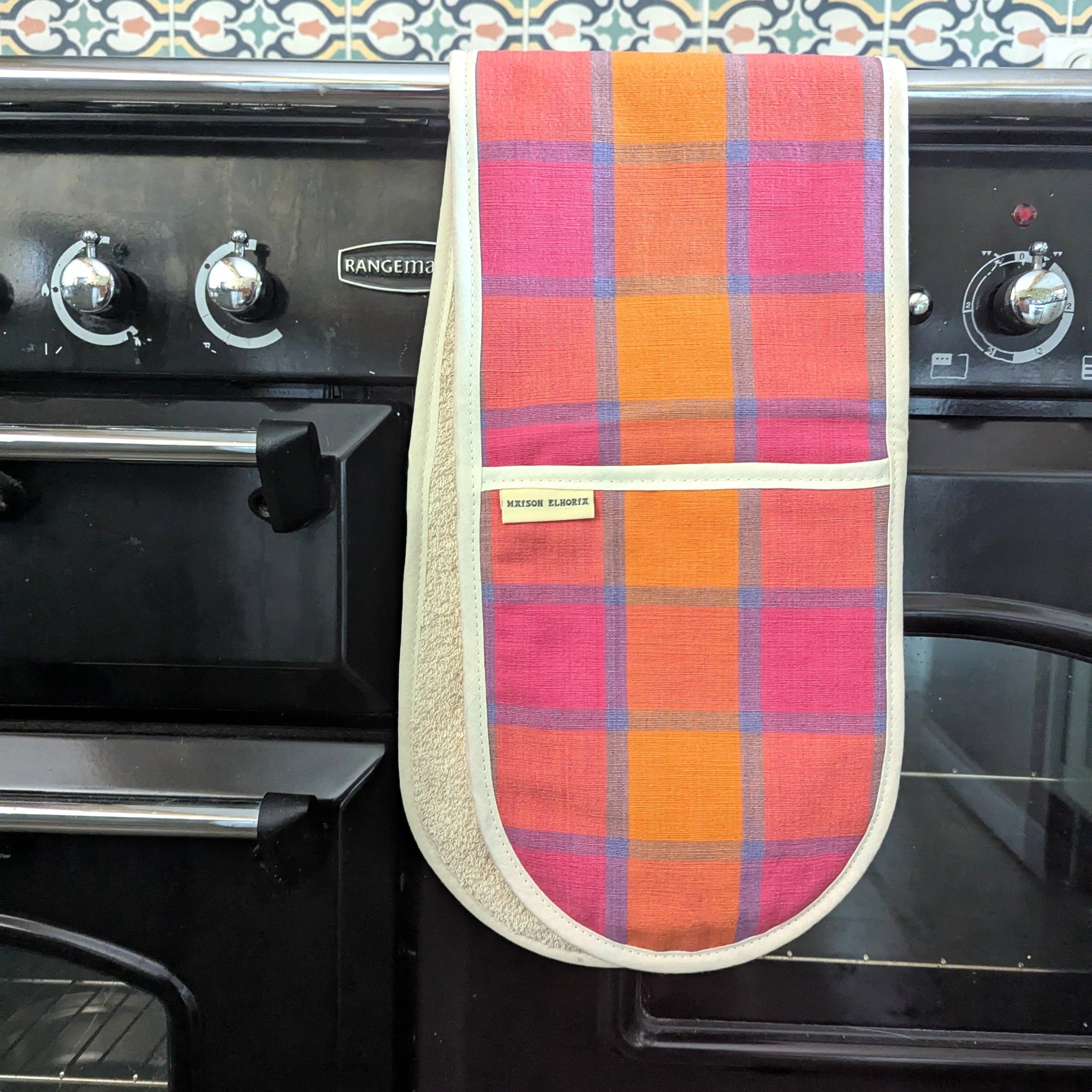 Multicolored checkered oven mitt hanging on a black oven with a patterned backsplash.