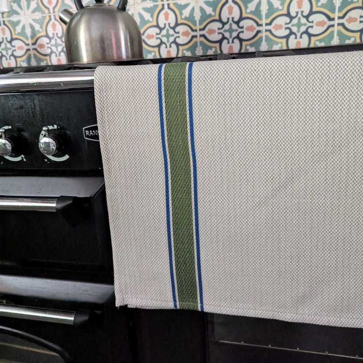 A kitchen towel with a textured cream weave hangs over a black stove, featuring a vertical stripe pattern of olive green, blue, and white. A silver kettle sits on the stove above, set against a background of teal and orange patterned ceramic tiles.