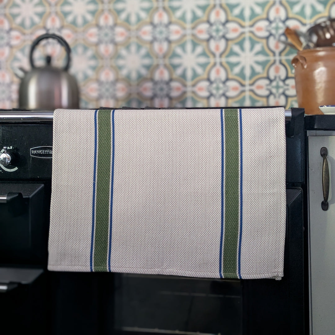 A kitchen towel with green and blue stripes hanging from the handle of a black oven in a kitchen setting with patterned wallpaper and a kettle in the background.
