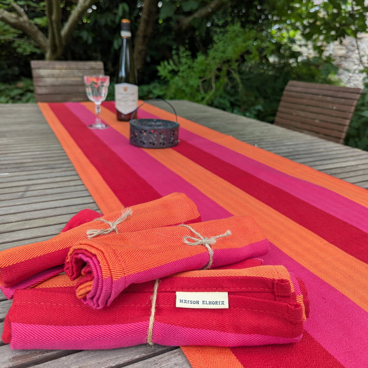Colourful striped table runner on a wooden table with folded red and pink cloths, glasses, and a bottle. Al fresco dinning