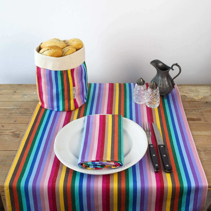 Table runner rainbow on a wooden table with a white plate , cutlery and a matching rainbow napkin and bread basket