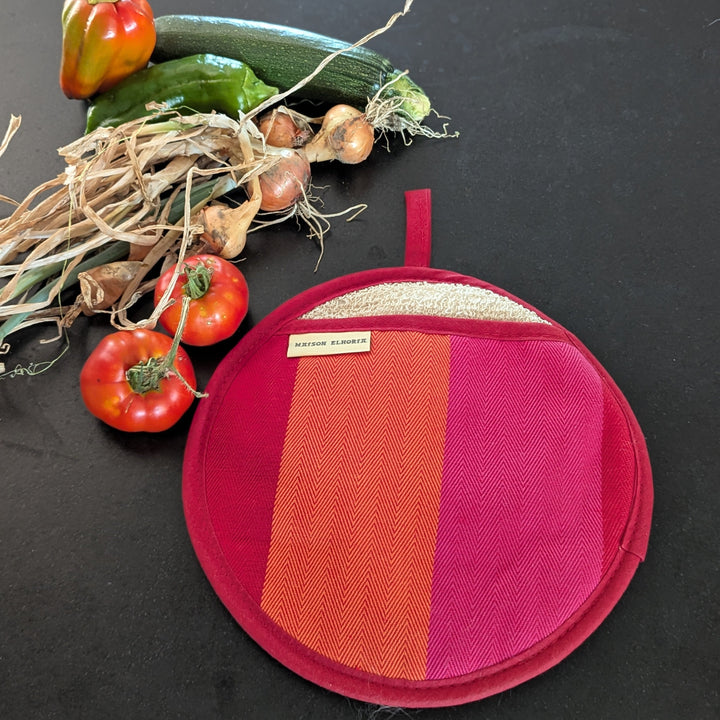 A round fabric potholder with a pink and orange striped design, displayed on a dark background next to fresh vegetables including tomatoes, garlic, and a zucchini.