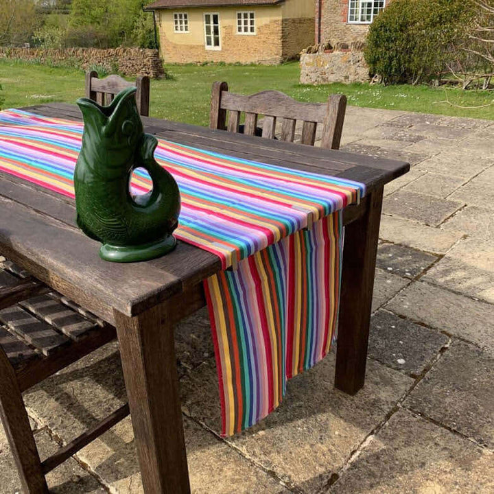 part cut image of Rainbow organic and recycled cotton table runner on a garden table outside with 4 chairs on a terrasse with grass and an old house  in the background a fish shaped vase on top 