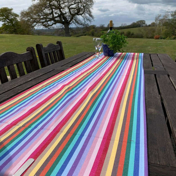 Rainbow organic and recycled cotton table runner on a garden table outside with 2 chairs  with grass and green hills and trees in the background and a plant pot 2 glasses and a bottle of wine  at the top far end of the table