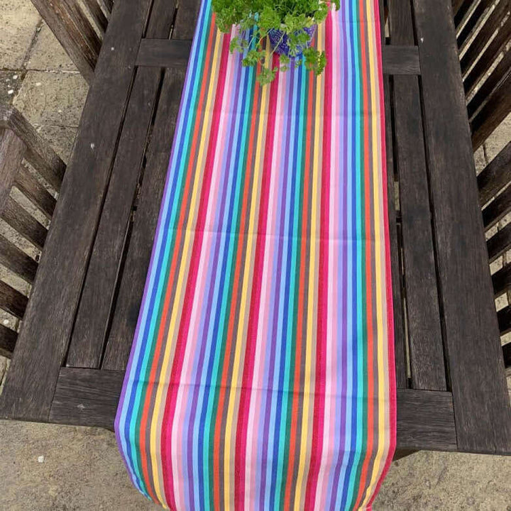 From above shot of a rainbow organic and recycled cotton table runner with a pot of parsley on top and 4 chairs on a garden table outside on a terrasse